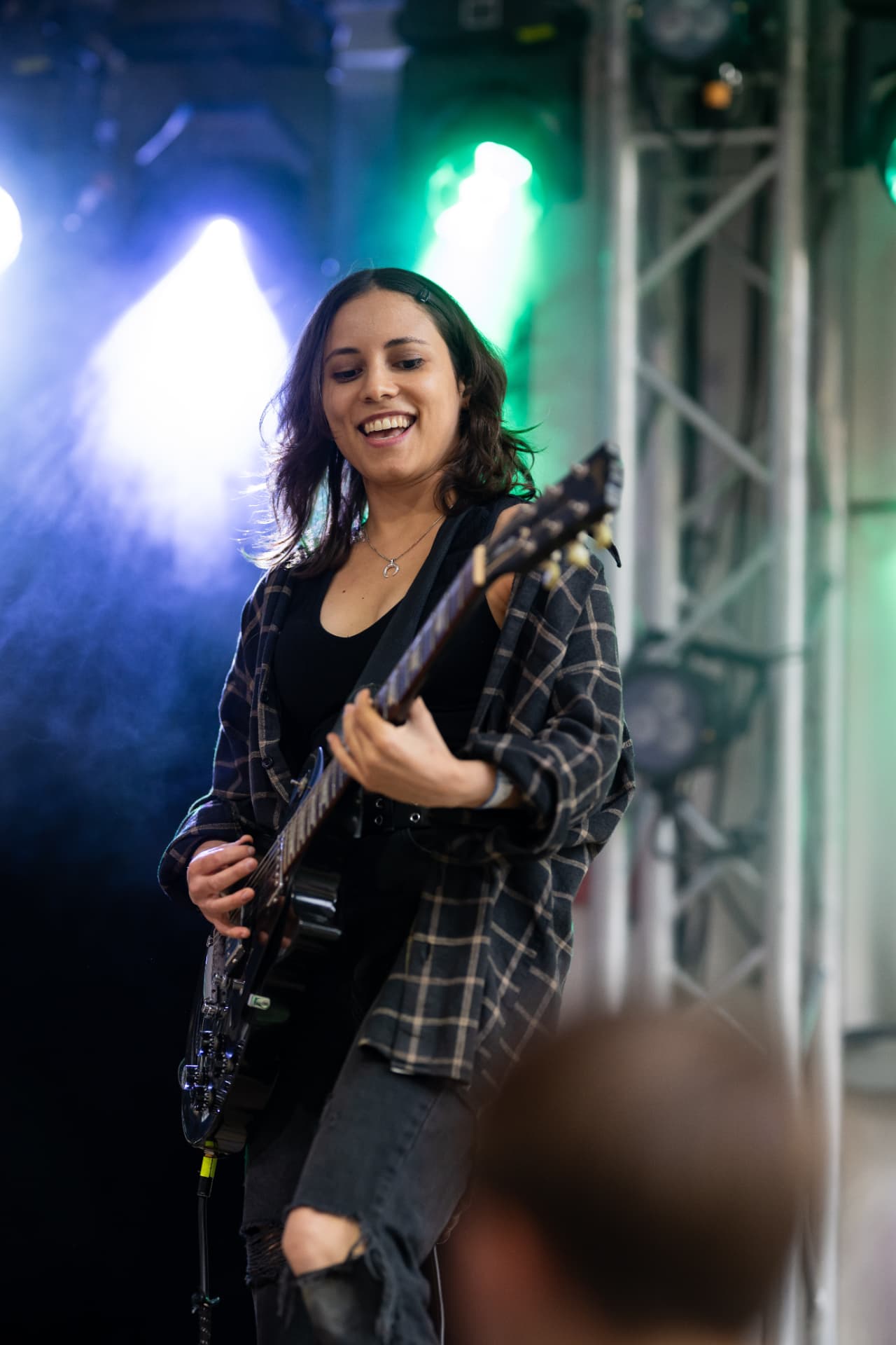 Gini Brown – Lächelnde Gitarristin mit kariertem Überhemd spielt schwarze E-Gitarre im blau-grünen Scheinwerferlicht