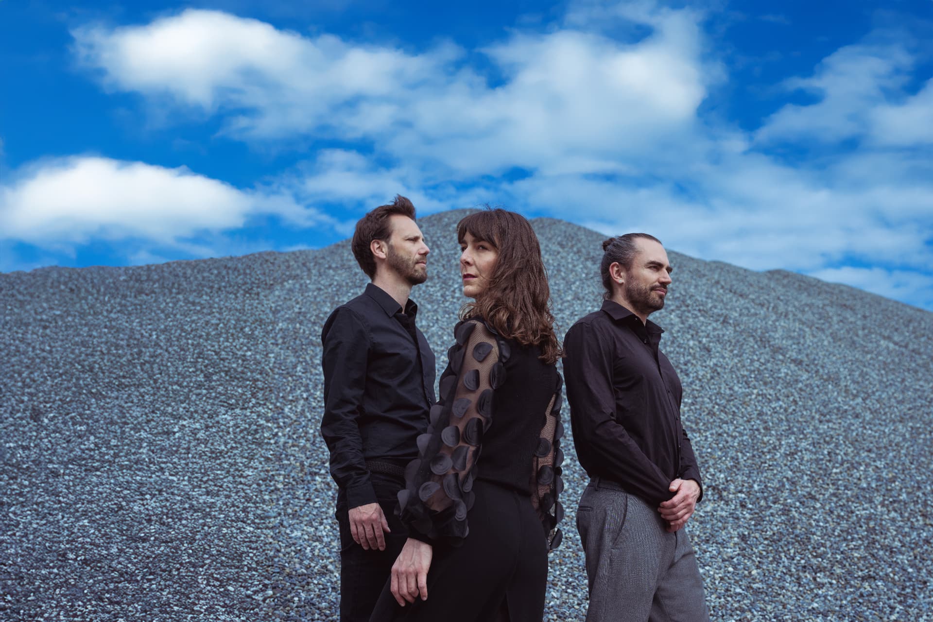 Die drei Musiker in Schwarz vor einem blaugrauen Schotterhügel unter bewölktem Himmel