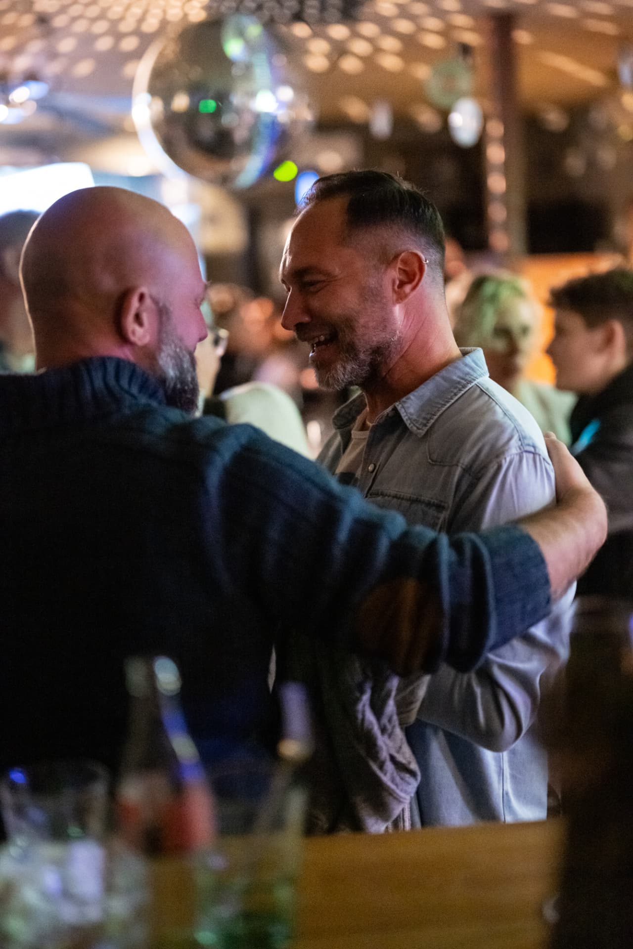 Two men embracing at a bar, one laughing, with other guests in the background