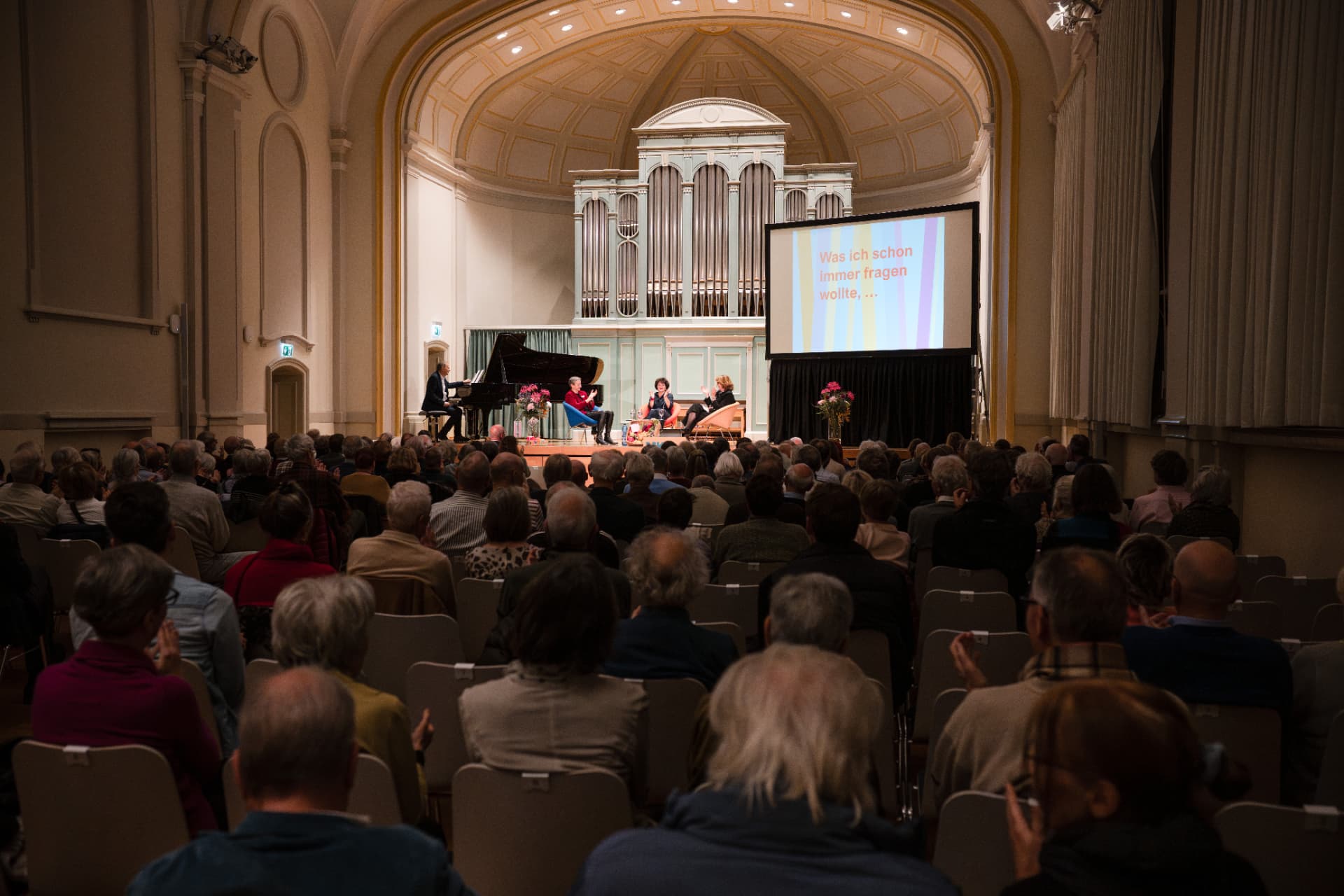 Totale des Festsaals: voll besetztes Publikum blickt zur beleuchteten Bühne mit Orgel und Leinwand