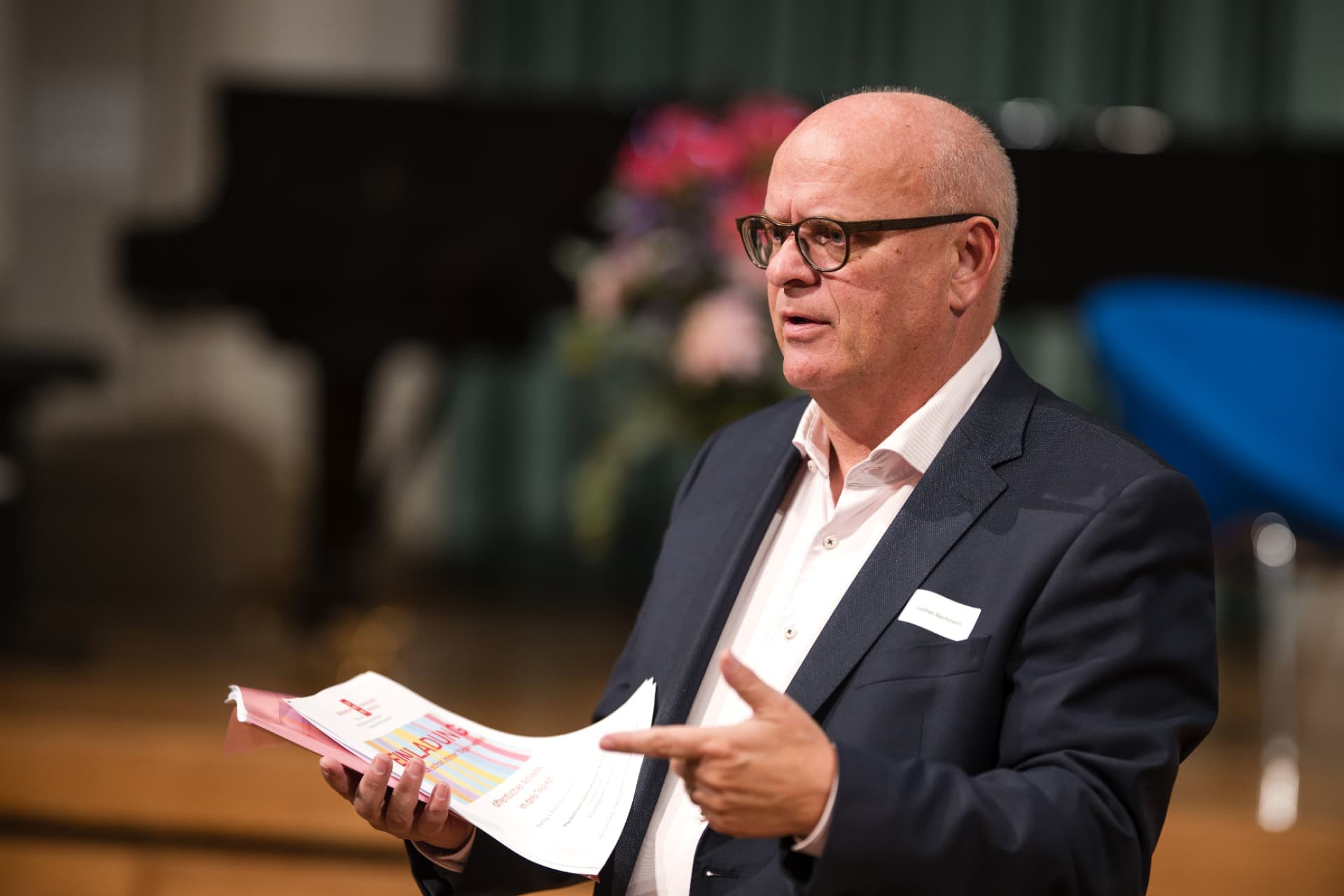 Speaker with glasses gesturing while holding a brochure