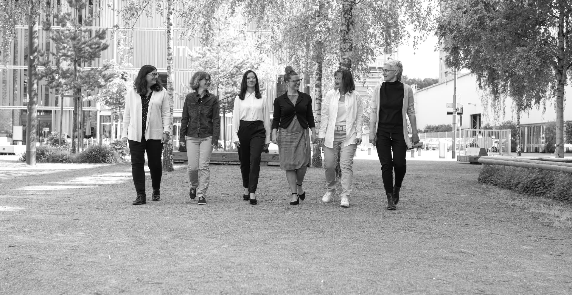 Sechs Frauen gehen lächelnd nebeneinander über einen Platz im Freien