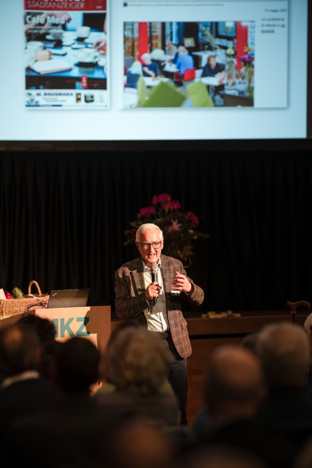 Older man speaking with a handheld microphone on stage, with a projection screen behind him