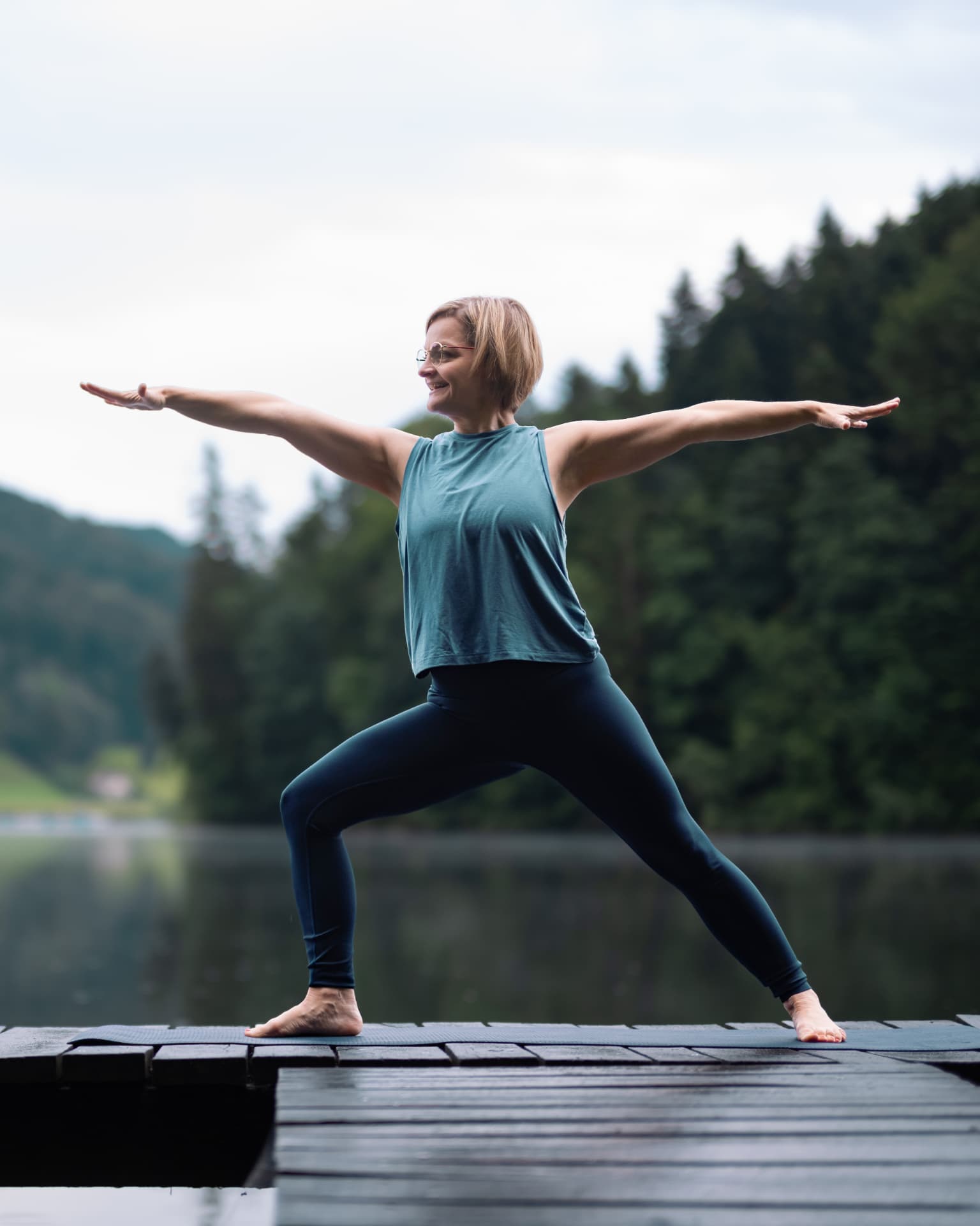Smiling woman in a yoga pose with outstretched arms on a wooden jetty by the lake