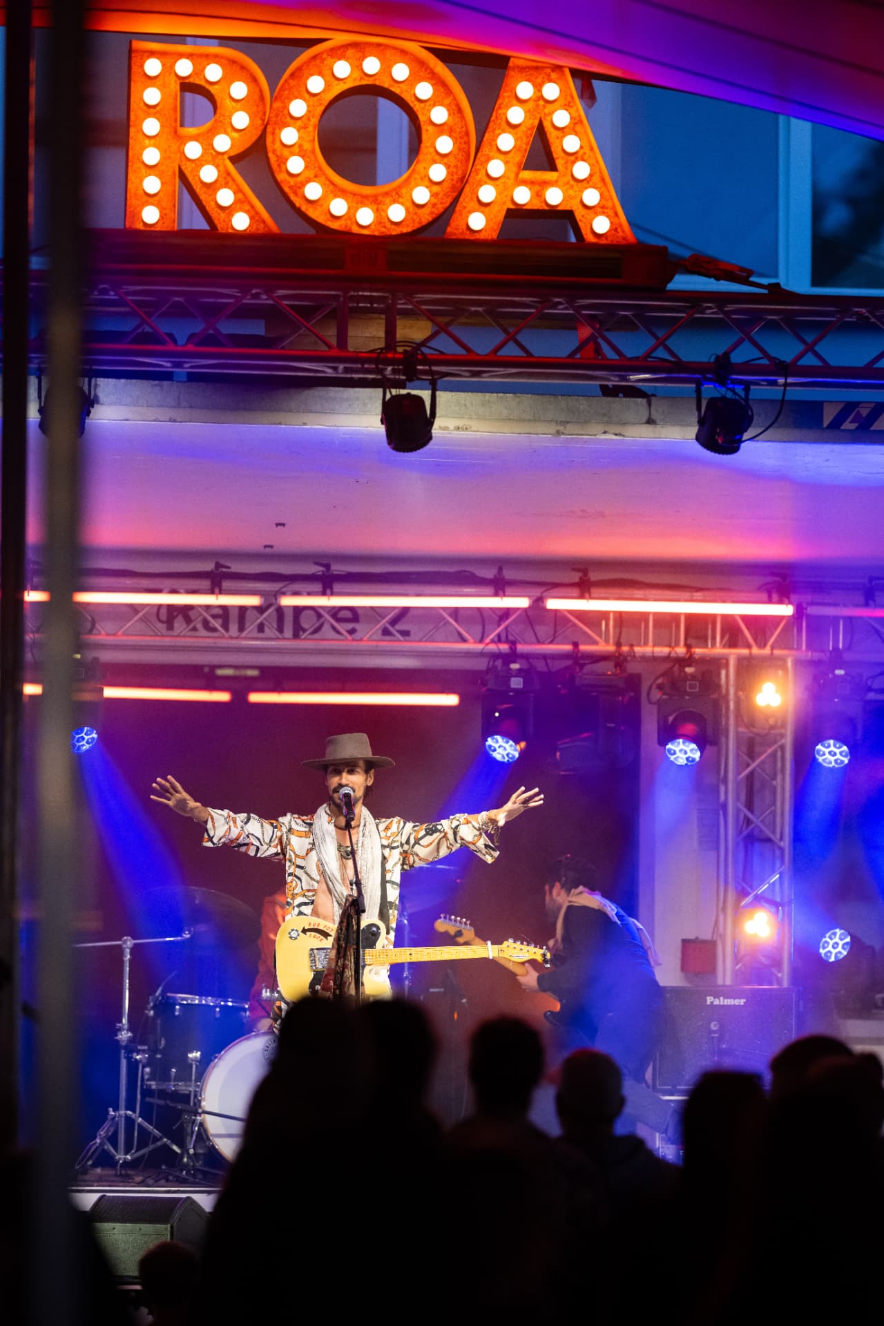 Captirando – Sänger mit Hut und Gitarre auf der Bühne, über ihm die beleuchteten Buchstaben ROA, davor Silhouetten des Publikums