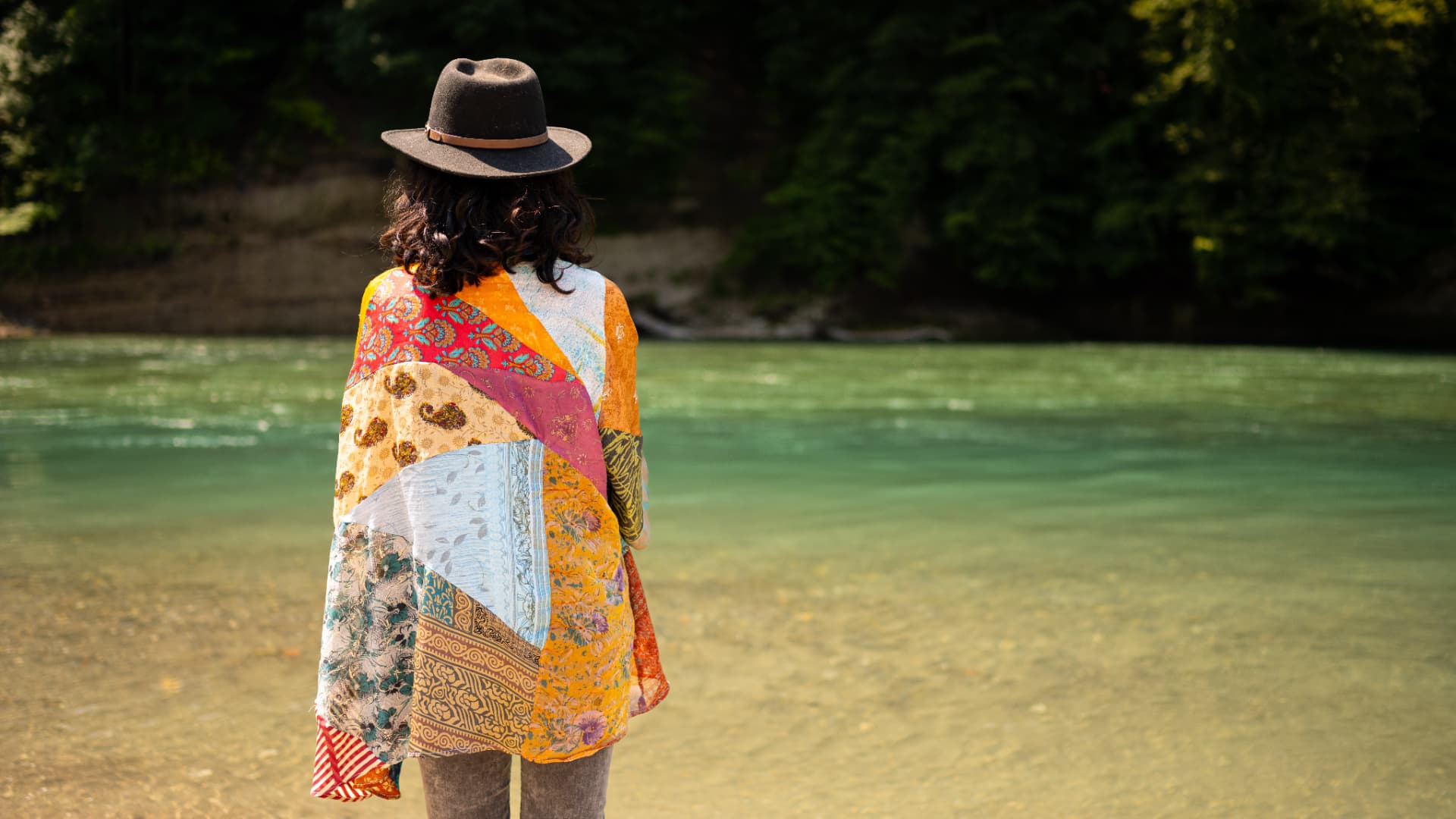 Meira Loom von hinten mit Hut und buntem Patchwork-Umhang, Blick auf das türkisgrüne Wasser