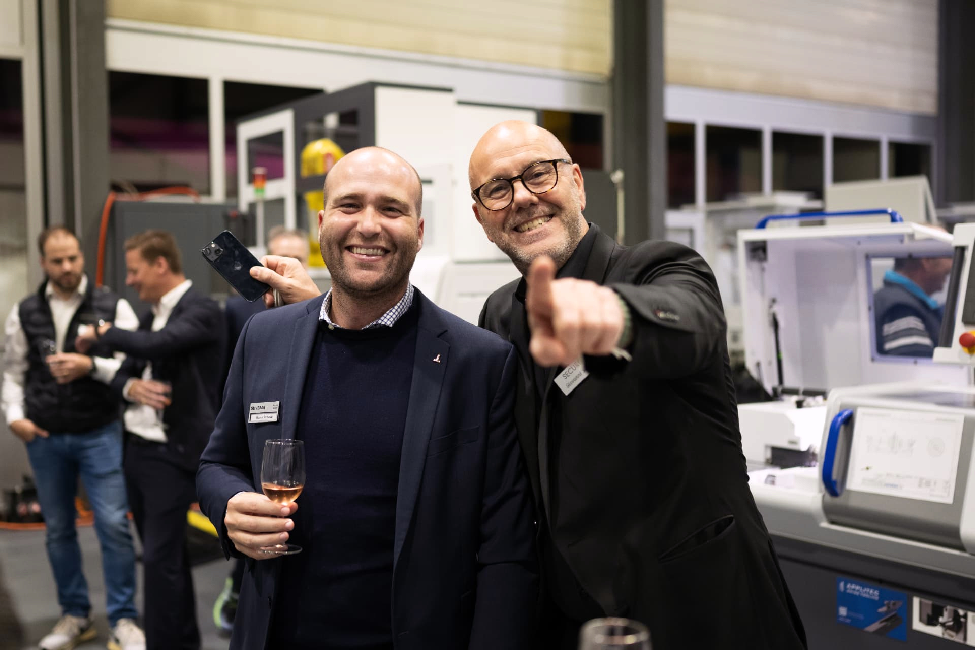 Zwei Männer lachen in die Kamera, einer deutet mit dem Finger in die Runde, dahinter CNC-Maschinen